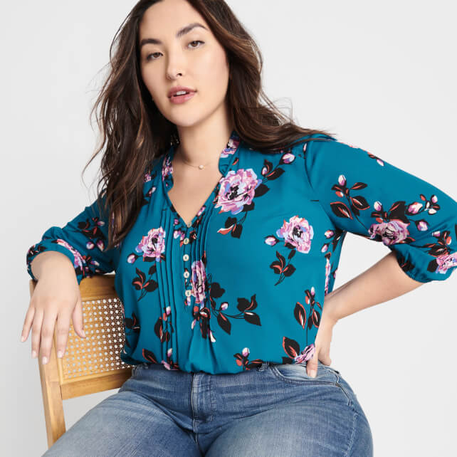 Plus model wearing teal blue floral top, blue jeans and sitting in a chair.